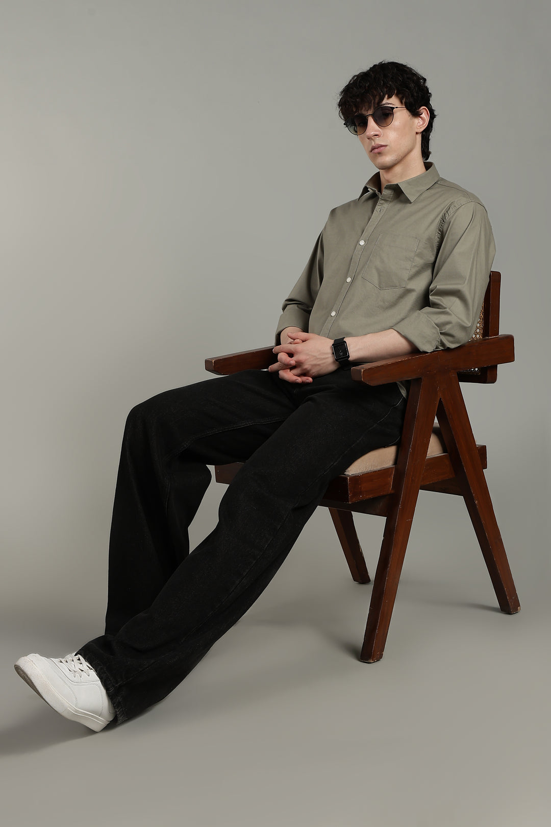 man sitting on a wooden chair wearing a sage green shirt and black pants against a gray background