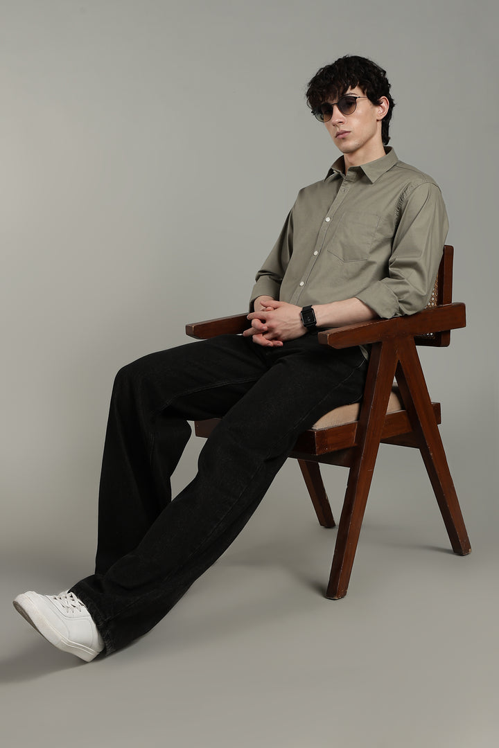 man sitting on a wooden chair wearing a sage green shirt and black pants against a gray background