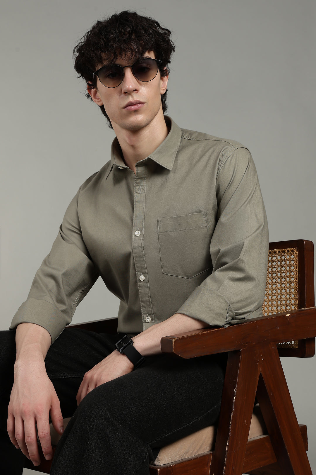 Man wearing sunglasses and a sage green shirt sitting on a wooden chair against a gray background