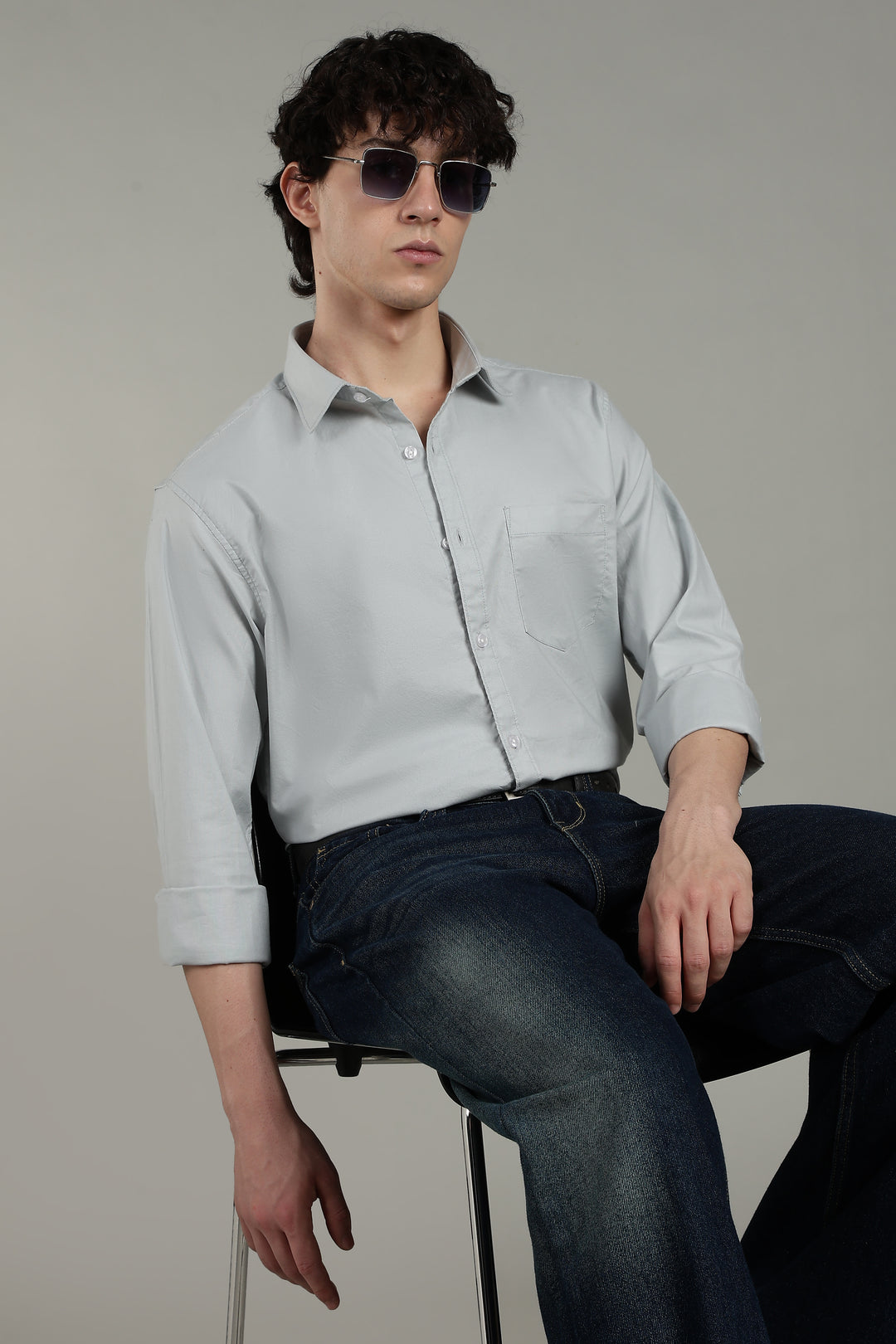 Man wearing a icy blue shirt and dark jeans sitting on a stool against a plain background