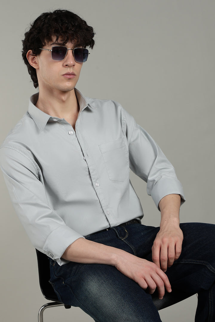 Man wearing a icy blue shirt and dark sunglasses sitting on a chair against a plain background