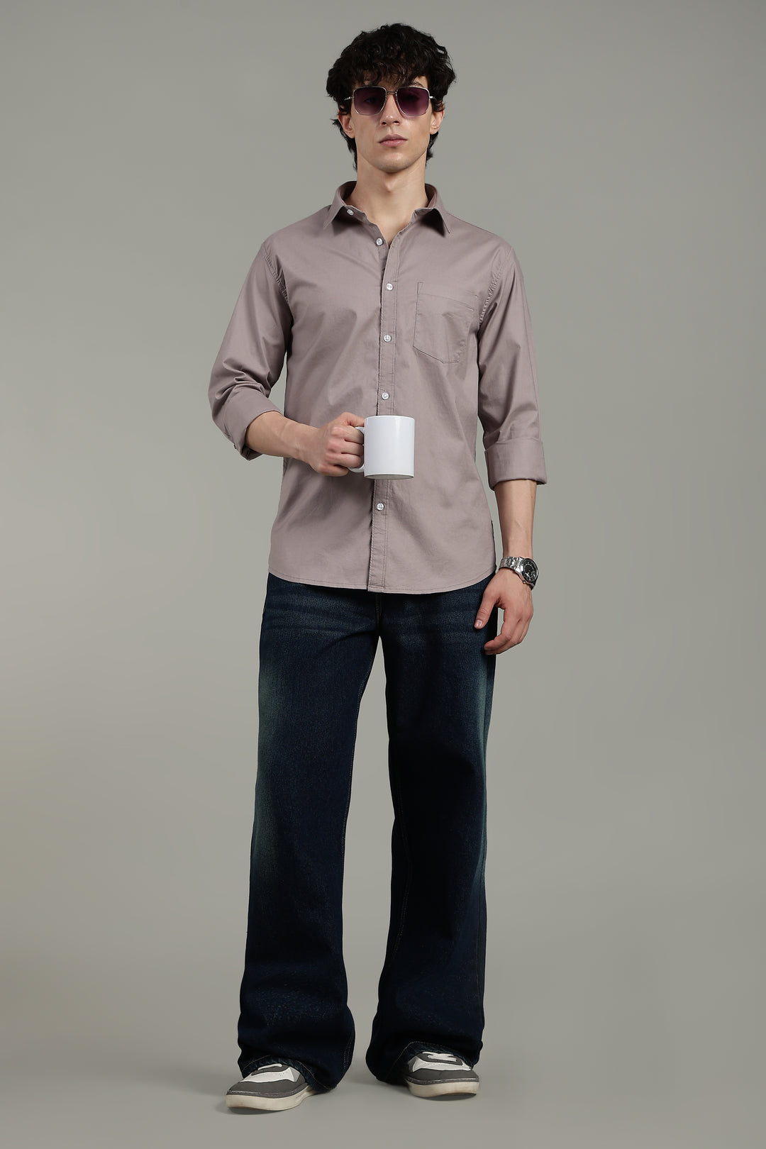 Man wearing a light mink shirt and dark jeans holding a white mug against a gray background