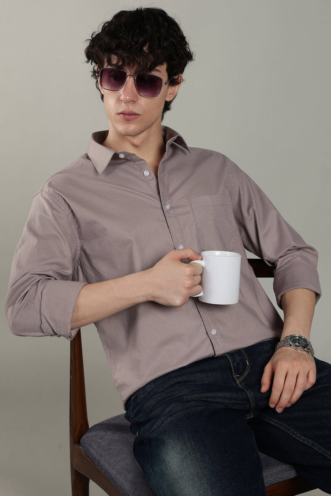 Man wearing sunglasses and a light mink shirt, holding a white mug, sitting on a chair against a plain background.