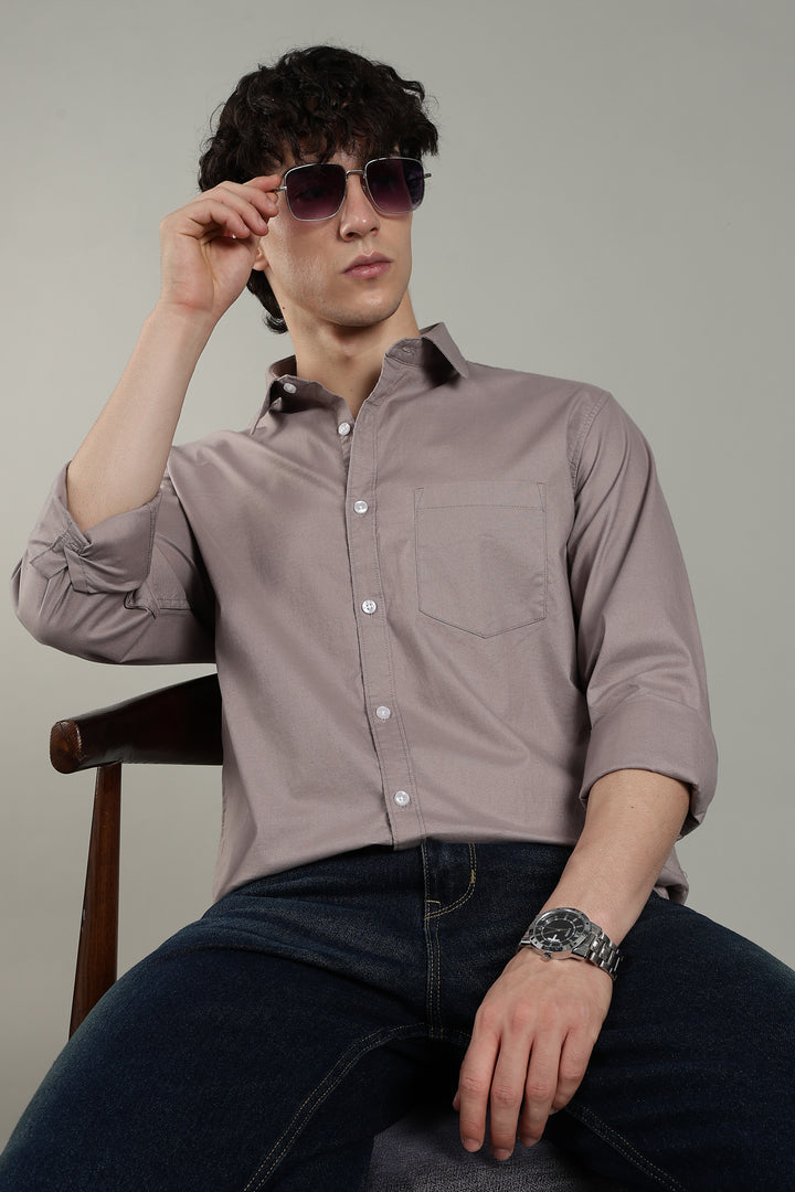 Man wearing a light mink button-up shirt and dark jeans, sitting on a chair against a plain background.