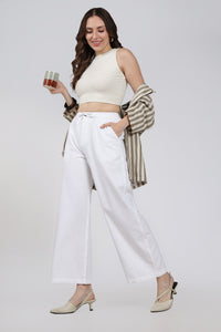 Woman in a cream top and white linen pants holding a coffee cup on a plain background