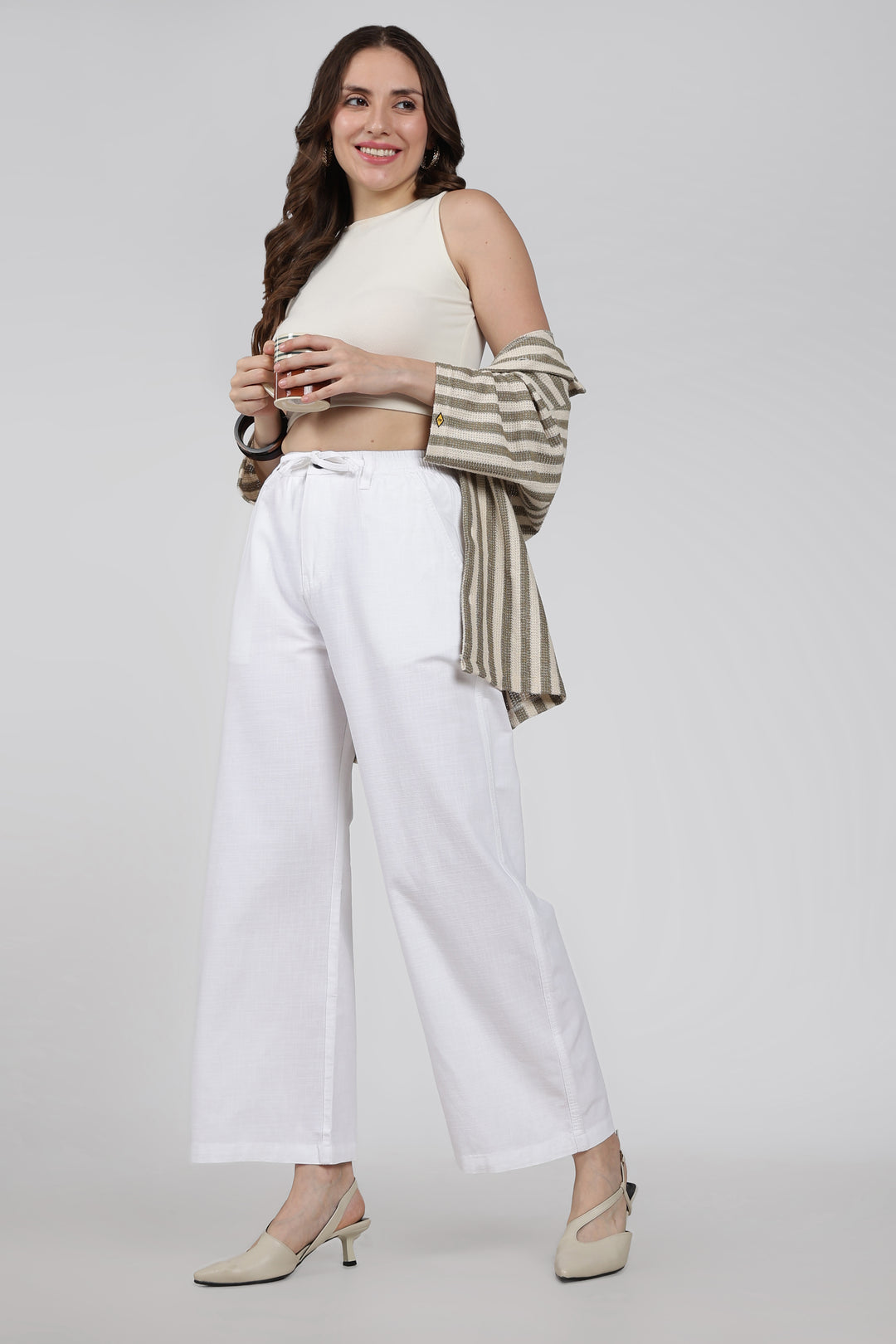 Woman wearing a cream top, striped long-sleeve shirt, white linen pants, and beige heels on a light gray background