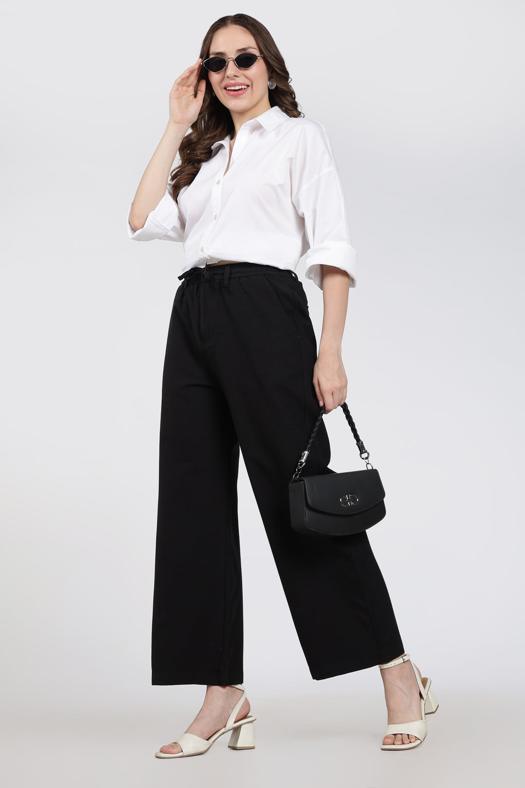 Woman wearing a white shirt and black linen pants, holding a black handbag on a white background