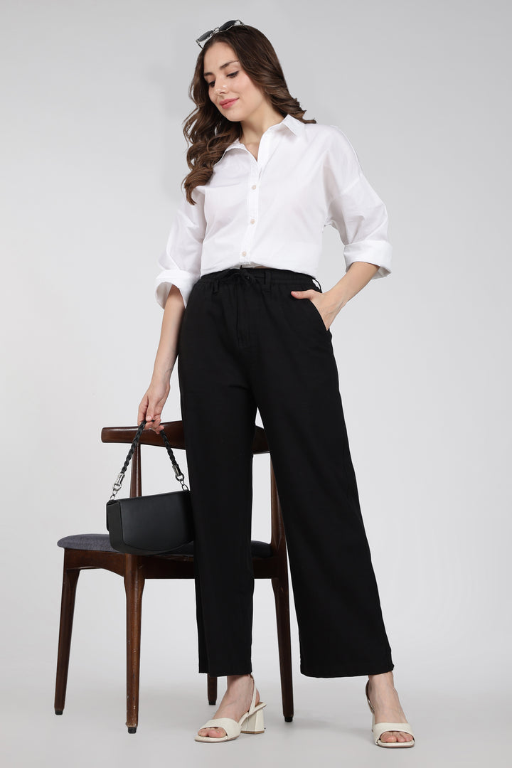 Woman wearing a white shirt and black linen pants, standing next to a wooden chair with a black handbag.