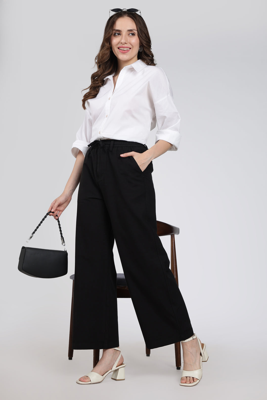 Woman wearing a white shirt and black linen pants with a black handbag on a gray background