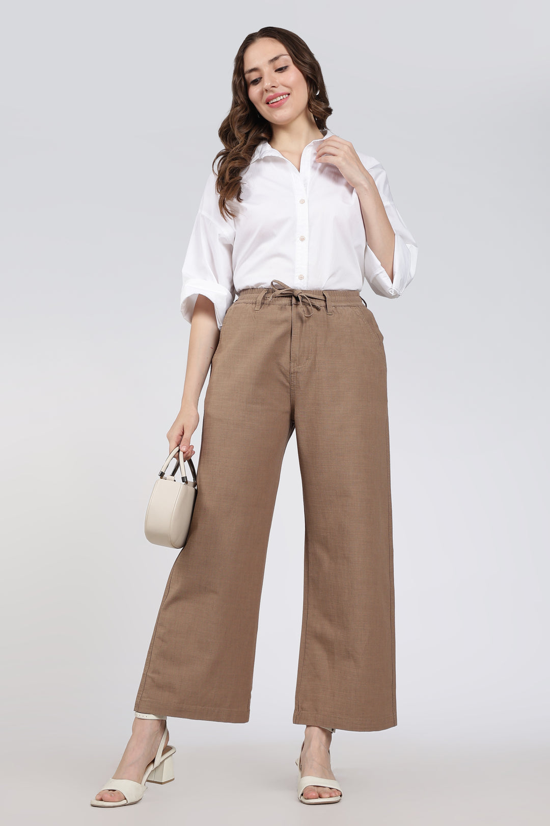 Woman wearing a white shirt and brown pants holding a beige handbag on a light gray background