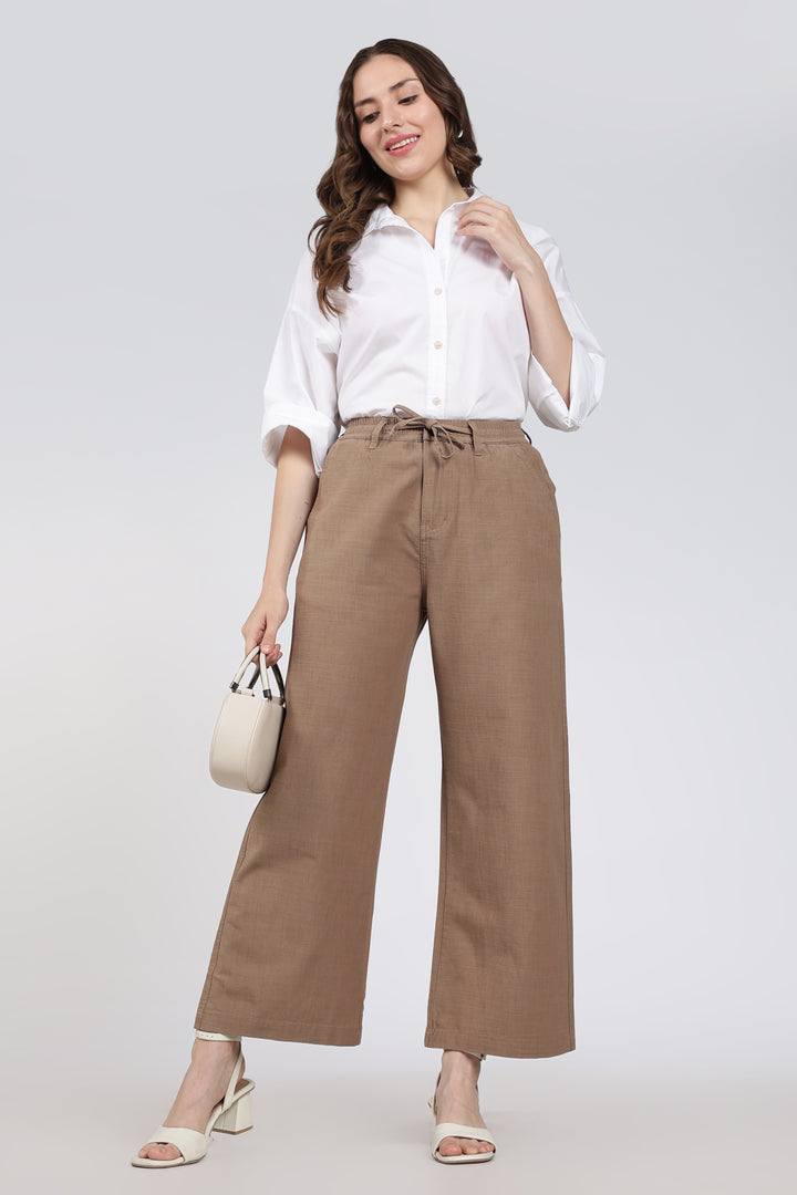 Woman wearing a white shirt and brown pants holding a beige handbag on a light gray background